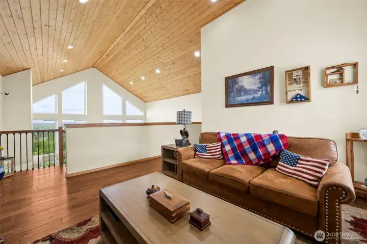 This upstairs bedroom at roughly 12 x 20 feet, is incredibly generous and absolutely drenched in natural light, while the jaw-dropping views looking out across the backyard deck toward the Olympic Mountains and even glimpses of Mount Rainier are amazing! Gorgeous hardwood floors continue throughout the upstairs level adding warmth and richness beneath the vaulted ceilings, while the handsome blue wall color gives the room a calm grounded feel that works beautifully with the natural wood details and surrounding scenery. A second window captures additional territorial views from another direction, the deep closet offers excellent storage behind a pine six-panel door and recessed lighting plus built-in ceiling speakers continue the thoughtful whole-home audio setup carried throughout the property.