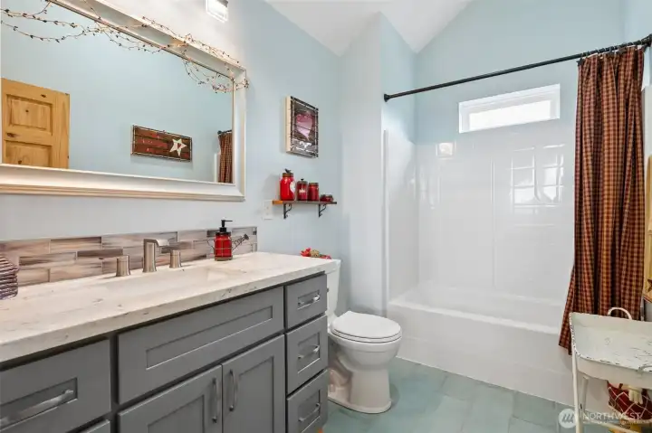 This upstairs bathroom feels bright, cheerful and surprisingly spacious thanks to the vaulted ceiling, soft blue wall color and natural light pouring in from the elevated privacy window above the shower. The full tub/shower combination, tile flooring and undermount sink vanity make the space highly functional for everyday living while still feeling cohesive with the warm custom finishes carried throughout the rest of the home. The elevated window placement is especially smart because it allows sunlight to flood the room while maintaining complete privacy, and the overall atmosphere feels fresh, calming and wonderfully comfortable for guests or upstairs living. It is also worth mentioning that this property includes two massive 3,000-gallon cistern holding tanks supplied from the well below the property, creating substantial water storage capacity that adds another layer of practicality and preparedness to this mountain retreat lifestyle. So yes, absolutely take the extra-long bath.