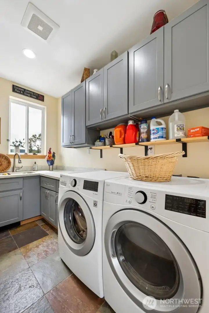 This laundry room is refreshingly generous in size and wonderfully practical, which feels exactly right for a custom home sitting on 20 acres because tiny cramped laundry spaces simply would not survive this lifestyle. Located conveniently on the main floor between the kitchen and foyer, the room functions beautifully for day-to-day living while still staying tucked out of the main entertaining spaces. At approximately 6.5 by 10 feet, there is ample room for the high-efficiency washer and dryer, folding space, cabinetry storage and a utility sink that becomes endlessly useful for everything from muddy boots to gardening cleanup to handwashing delicate. The window keeps the room bright and welcoming instead of cave-like, while the pocket door helps contain noise and keeps the overall flow of the house feeling clean and uninterrupted.