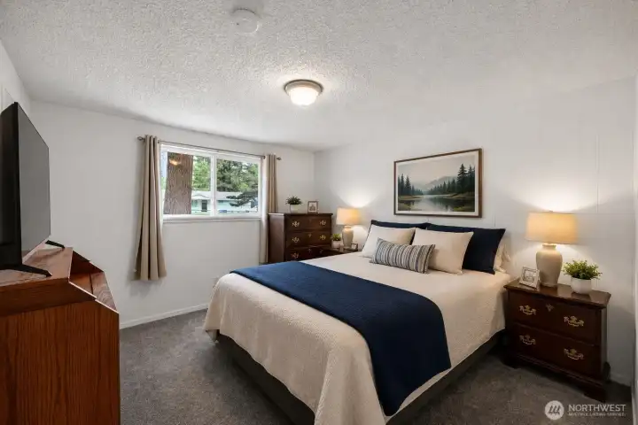 Comfort and simplicity come together in this inviting bedroom retreat. Soft natural light, neutral tones, and warm wood furnishings create a relaxing atmosphere, while the spacious layout easily accommodates a king-sized bed. The large window brings in views of mature greenery outside, adding to the room’s peaceful feel. A comfortable and versatile space designed for everyday living and restful evenings.