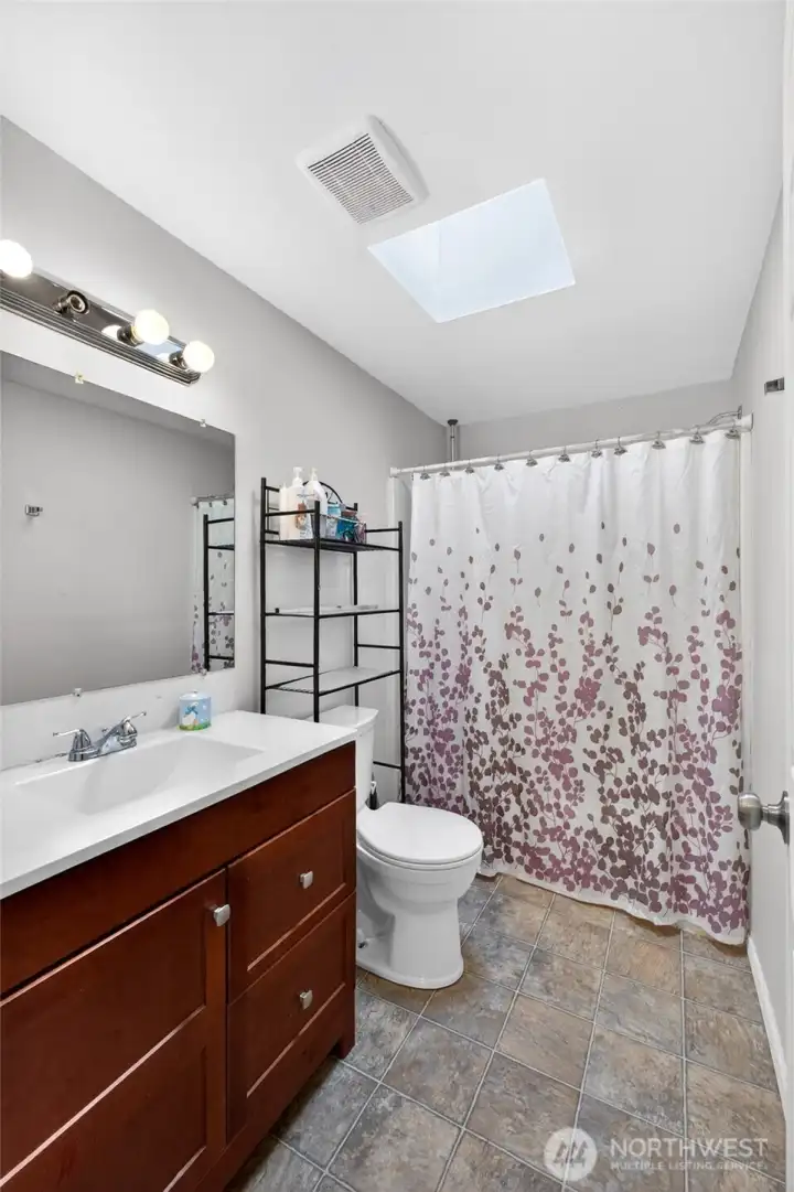 Clean and functional full bathroom featuring an updated vanity with rich wood cabinetry, modern fixtures, and durable tile flooring. The skylight adds natural brightness to the space, creating an open and airy feel, while the neutral finishes make it easy to personalize to your style.