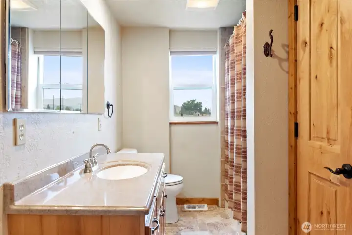 Bright bathroom with a solid wood door, mirrored storage, and a tiled shower/tub combo. Natural light from dual windows adds warmth and charm.
