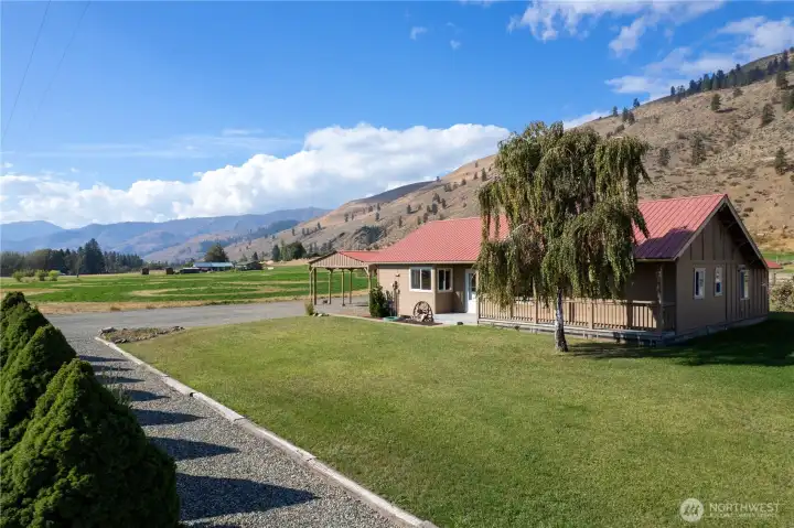 Beautiful side view of the home with covered porch, established trees, and wide-open green space leading to mountain views.