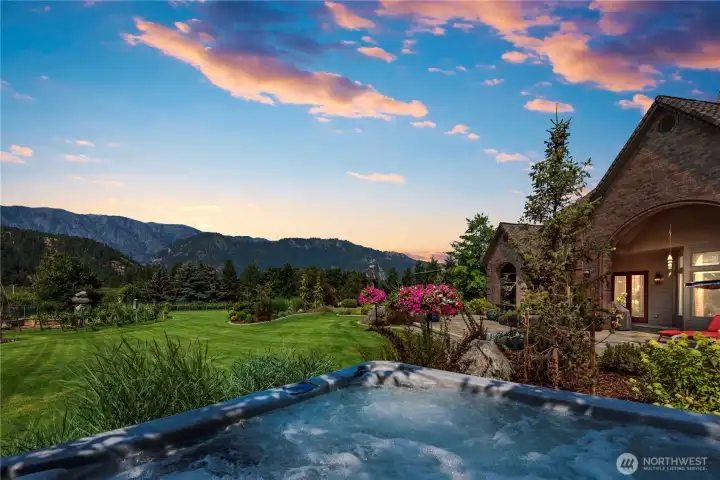 Beautiful outdoor living space featuring a hot tub, manicured grounds, and breathtaking sunset views across the valley toward the Cascade Mountains and Enchantment Range.