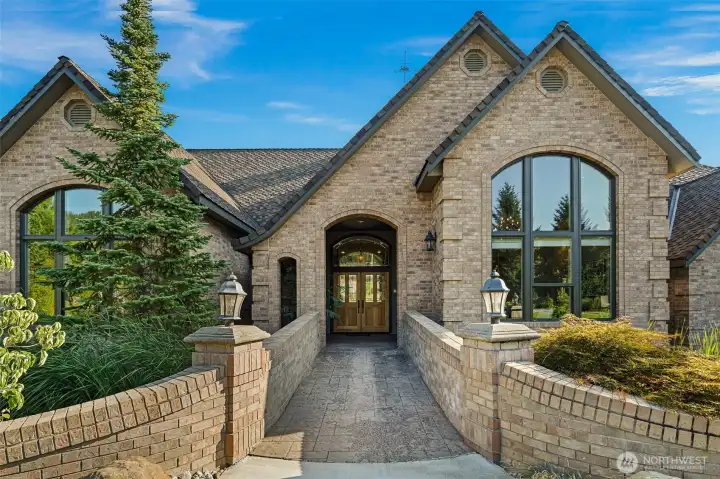 Striking brick façade welcomes you with a dramatic arched entry, elevated walkway with brick balustrades, and rich wood double doors framed by soaring windows that create a warm, inviting arrival.
