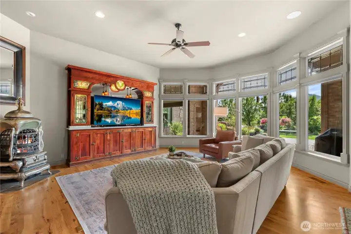 Light-filled living room featuring expansive windows with transom accents that frame beautiful outdoor views. Warm hardwood floors, high ceilings, and a cozy stove create an inviting space for relaxing or entertaining.