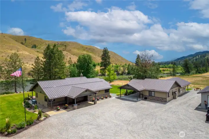 A stunning riverside property featuring two beautifully designed single-level homes with modern metal roofs and stone accents. Nestled against rolling hills and lush greenery, the residences are connected by concrete sidewalks and surrounded by an expansive gravel courtyard. The serene landscape, complete with mature trees and a nearby river, creates the perfect blend of comfort, privacy, and natural beauty.