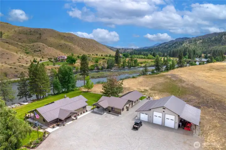 This aerial view highlights the property’s full layout, including two residences, a large shop/garage, and direct river frontage. Surrounded by rolling hills and open landscape, the estate offers both privacy and room for recreation.