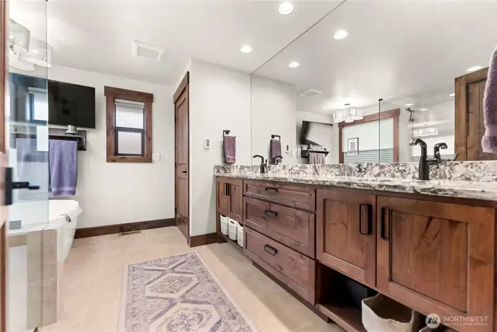 Adjacent to the tub is a spacious double vanity with rich wood cabinetry, granite countertops, and undermount sinks, complemented by sleek black faucets and fixtures. The vanity area also includes a large wall mirror, ample drawer and shelf storage with woven baskets, and dual towel racks.