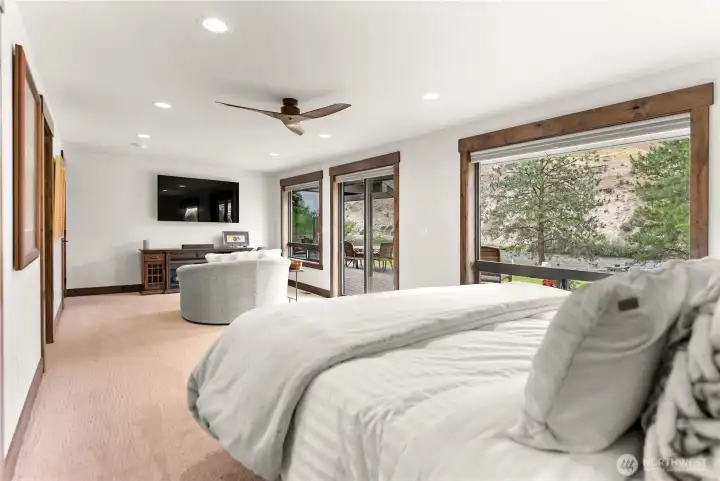 This cozy corner of the primary bedroom doubles as a private entertainment area, complete with a wall-mounted flat-screen TV and an electric fireplace set into a wooden media console. The oversized round chair remains the focal point of comfort, while two sets of sliding doors open to a lush green yard and patio seating area, offering stunning views and easy indoor-outdoor living.
