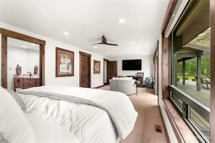 A view from inside the primary bedroom highlights its warm and inviting design, featuring rich wood trim, doors, and baseboards that add a rustic elegance. The king-sized bed is complemented by simple yet tasteful furnishings, including a vintage-style lamp and a wooden trunk at the foot of the bed. Artwork along the wall and recessed lighting create a cozy, lived-in ambiance, while the open floor plan enhances the spacious feel.
