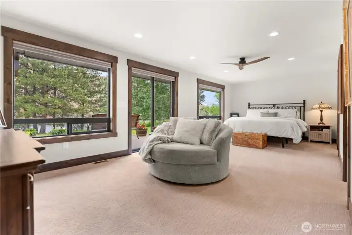 This spacious primary bedroom offers a serene retreat with expansive windows that frame picturesque views of the surrounding trees and landscape. The room is filled with natural light, and features a cozy reading nook with a plush round swivel chair, perfect for relaxing. Sliding glass doors provide direct access to a private outdoor patio area, seamlessly blending indoor comfort with outdoor tranquility.