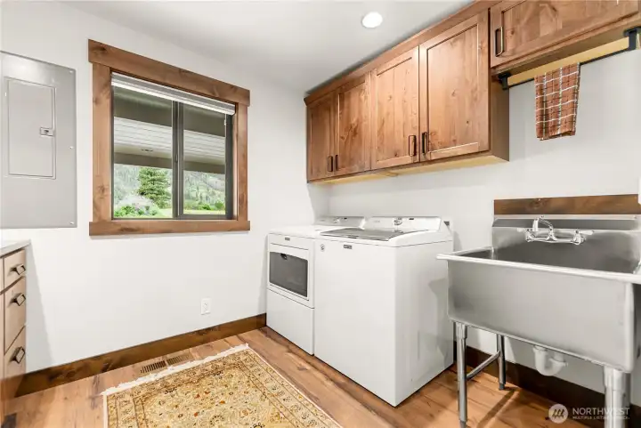 Bright and functional laundry room featuring modern Maytag washer and dryer, stainless steel utility sink, and abundant cabinetry for organization. Large window brings in natural light and frames peaceful mountain views, making everyday chores more enjoyable.