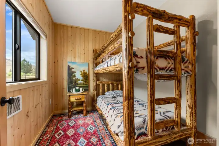 A cozy bunk room with warm wood paneling, rustic log-frame bunks, and a colorful accent rug — perfect for hosting family or guests in comfort and style.