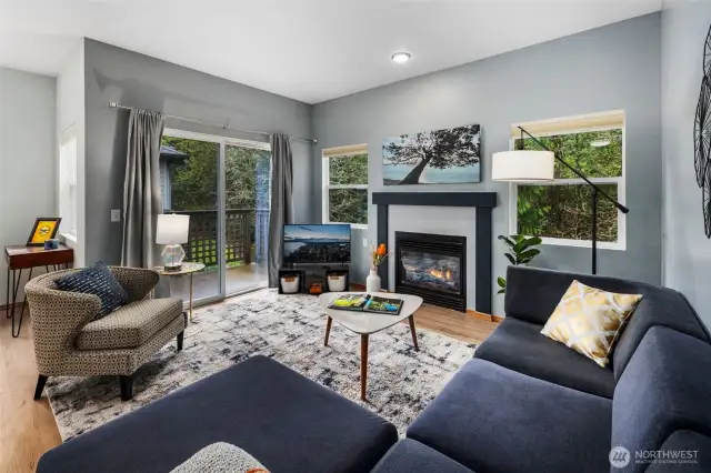 Bright and welcoming living room centered around a cozy gas fireplace—perfect for relaxing or entertaining. Large windows and sliding doors open to a private deck surrounded by lush green trees, blending indoor comfort with outdoor serenity