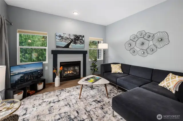 Bright and welcoming living room centered around a cozy gas fireplace—perfect for relaxing or entertaining. Large windows and sliding doors open to a private deck surrounded by lush green trees, blending indoor comfort with outdoor serenity