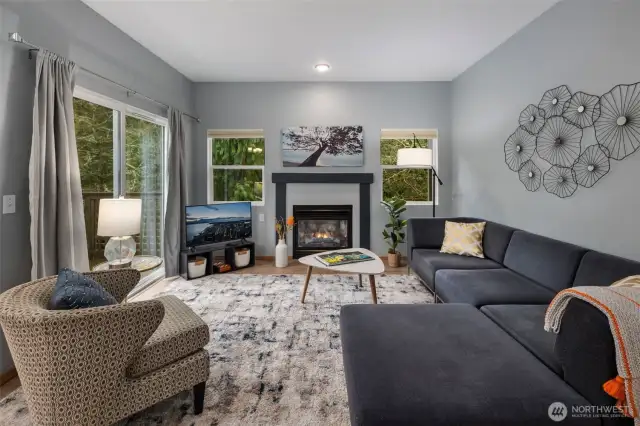 Bright and welcoming living room centered around a cozy gas fireplace—perfect for relaxing or entertaining. Large windows and sliding doors open to a private deck surrounded by lush green trees, blending indoor comfort with outdoor serenity