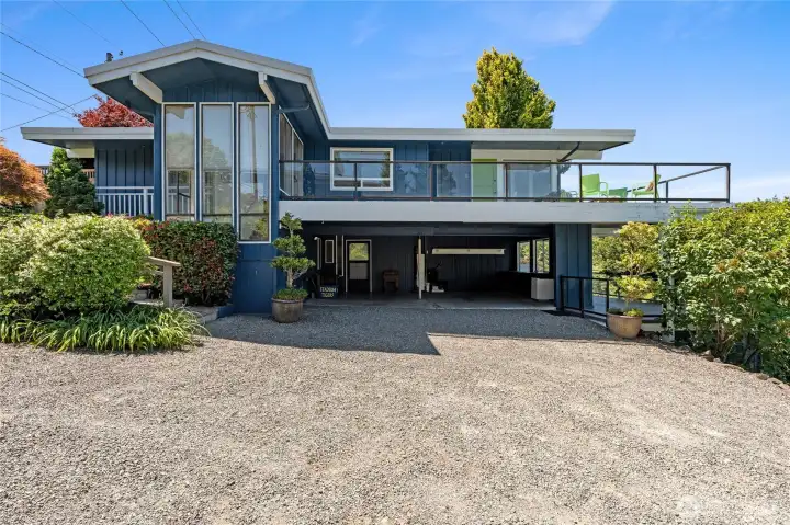 Side Elevation of the home showcasing two large exterior decks with glass railings. Tall windows on three sides of the staircase.  Upper wraparound deck off of the great room & lower deck with Hot Springs Spa.  Two car parking in the lower level for easy access to the home & a detached garage on the lower level.