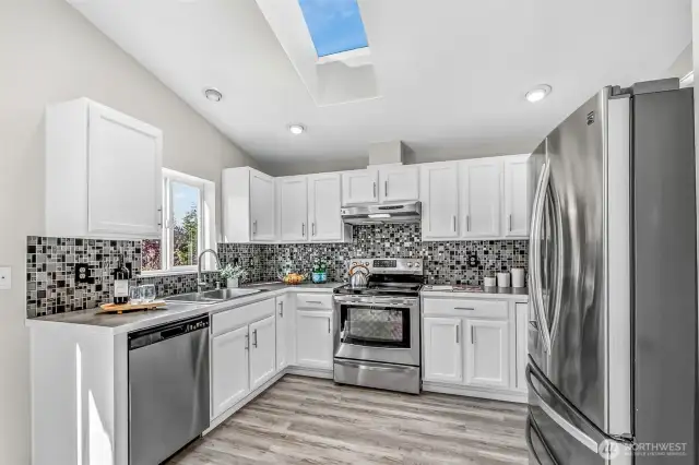 Crisp white cabinetry, mosaic tile backsplash, and stainless appliances define the kitchen. A skylight and window over the sink bathe the workspaces in natural light, while new LVP flooring offers durability and style.