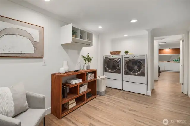 This basement laundry area features side-by-side front-loading washer and dryer units, known for their energy efficiency and gentle handling of clothes. The side-by-side arrangement offers convenient access to both machines, making laundry tasks more manageable. Plenty of shelf space for detergents, a hanging rack, and room for a large table for folding.