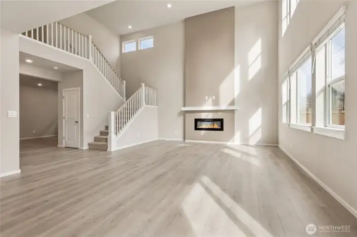 Stunning wide-angle view of the great room featuring soaring two-story ceilings, an open staircase with white painted wood railing, a modern linear electric fireplace, and oversized windows bathing the space in beautiful natural light. Photos are representative only and may display different color package and/or upgrades.