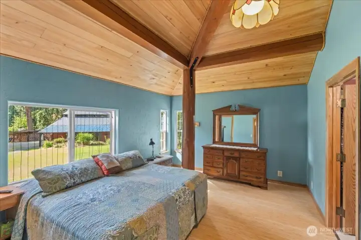 Spacious primary bedroom with vaulted wood ceiling, exposed timber accents, and large picture window overlooking the grounds. Natural light and warm wood details create a comfortable retreat. Spacious primary bedroom with vaulted wood ceiling, exposed timber accents, and large picture window overlooking the grounds. Natural light and warm wood details create a comfortable retreat.