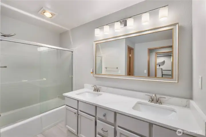 Sleek lower level updated bathroom featuring double vanity, quartz counters, framed mirror, modern lighting, glass shower enclosure, and clean contemporary finishes.