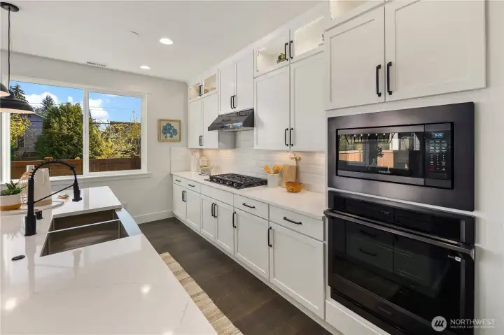 Kitchen  Full-height white cabinetry and quartz surfaces define this open concept kitchen, designed for modern Pacific Northwest living in Bothell’s Canyon Park area. A generous island with integrated sink anchors the space, complemented by a gas cooktop, wall oven, built-in microwave, and under-cabinet lighting. Expansive rear windows invite natural light while wide plank flooring adds warmth and contrast. The layout supports seamless flow between cooking and gathering, reflecting the elevated finishes expected in luxury new construction within the Northshore School District.    These photos are from a past Robinson Plan at a different community, so overall plan, colors, building orientation, finishes, home outlook, and other details can and WILL vary. All photos, renderings, site maps, and floorplans are for visualization purposes only.