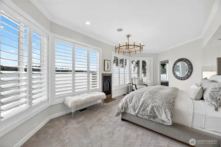 The serene primary suite offers lake views through plantation-shuttered bay windows, a cozy fireplace, and a chandelier that brings quiet elegance to every morning.