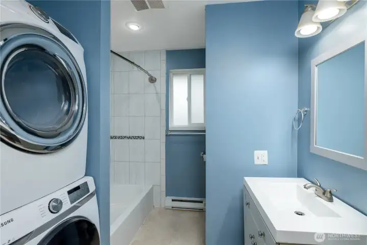 This bright and functional bathroom features a sleek vanity with brushed nickel fixtures, a well-lit mirror, and fresh blue walls that create a calming atmosphere. A convenient stacked Samsung washer and dryer maximize efficiency, making this bathroom a perfect blend of utility and comfort.