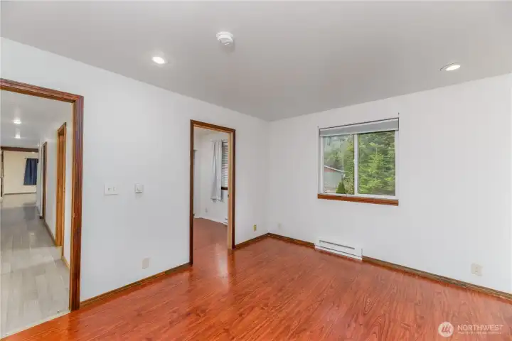 Another view of the primary bedroom showcasing its bright window, recessed lighting, and rich flooring. This angle highlights the functional layout with direct access to a secondary bedroom or office space, offering flexibility for a home office, nursery, or private retreat. The hallway connection creates a natural flow into the rest of the home.