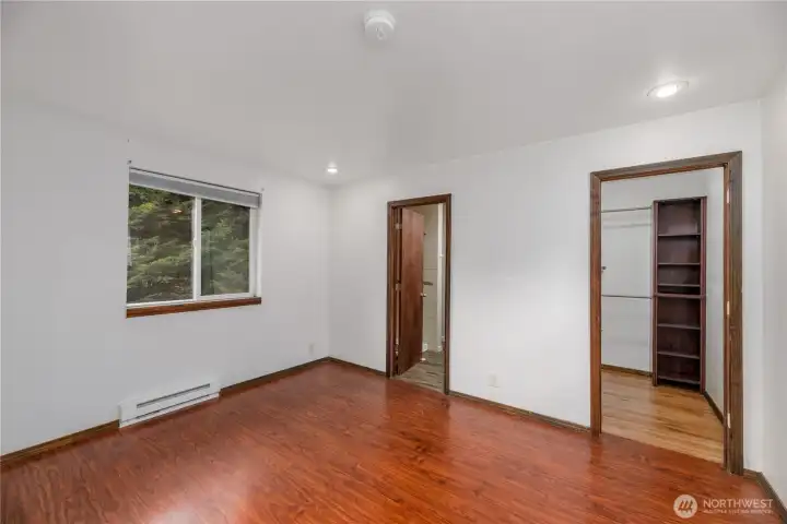 Inviting bedroom featuring rich wood-look flooring, recessed lighting, and a large window framing natural outdoor views. The room includes a walk-in closet with built-in shelving for organized storage and direct access to a private bathroom, making it an ideal primary suite. Neutral walls and trim provide a versatile backdrop, ready for personal design touches.