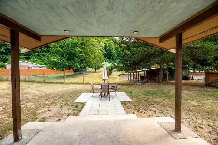 Covered patio overlooking a spacious, fully fenced yard with mature trees for added privacy. A paved patio area provides the perfect spot for outdoor dining and entertaining, complete with space for a table and chairs. The large open yard offers plenty of potential for gardening, play, or pets, making it a versatile extension of the home’s living space.