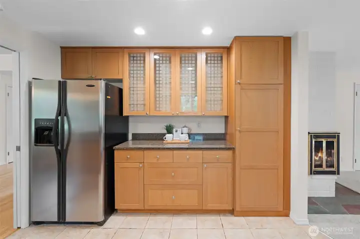 Dedicated storage wall with custom cabinetry, glass-front uppers, and extended counter space for prep or serving. Adjacent layout flows easily toward the main living area with fireplace nearby.