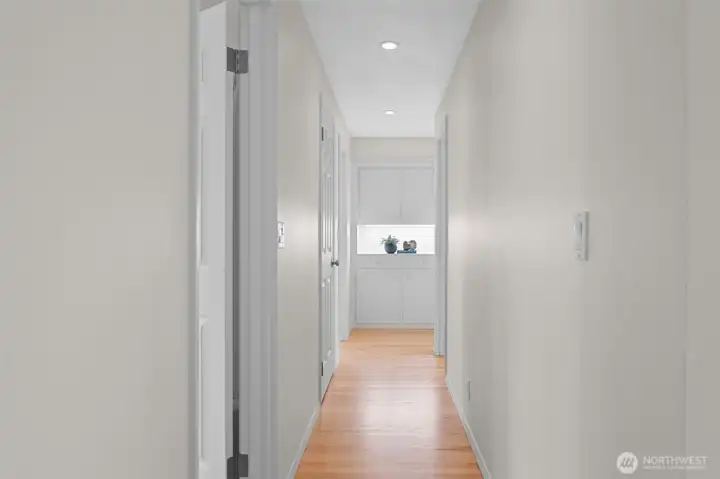Hallway with clean lines, wood flooring, and recessed lighting leading to built-in storage at the end. Thoughtful layout places the primary suite and two additional bedrooms together for a connected and functional design.