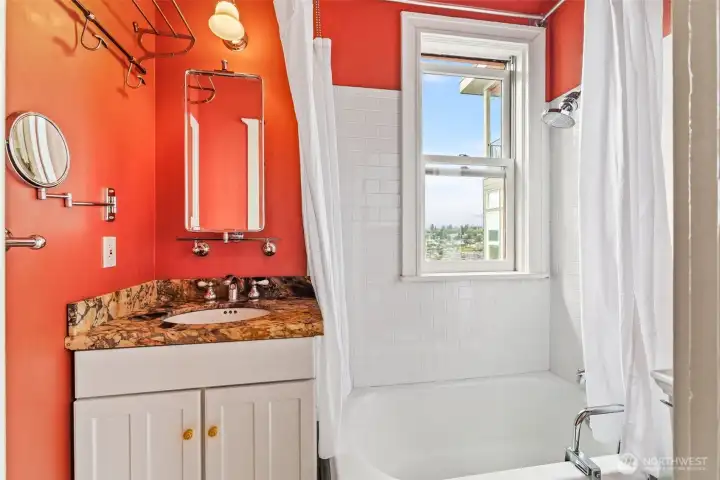 Bathroom with Character – Features a bold red accent wall, granite countertop, and classic tile shower surround with a window offering a view.