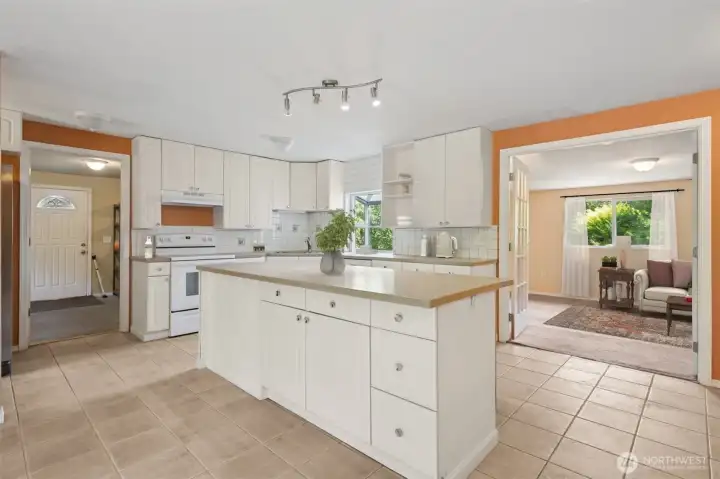 The kitchen keeps it classic with white shaker-style cabinets, silver hardware, and a clean tile backsplash. White appliances blend in, while a corner sink sits under a garden shelf window that’s basically begging for fresh herbs. The back door carries the same arched glass detail as the front, and through the French doors you’ve got flex space that works as a third bedroom or whatever you need.