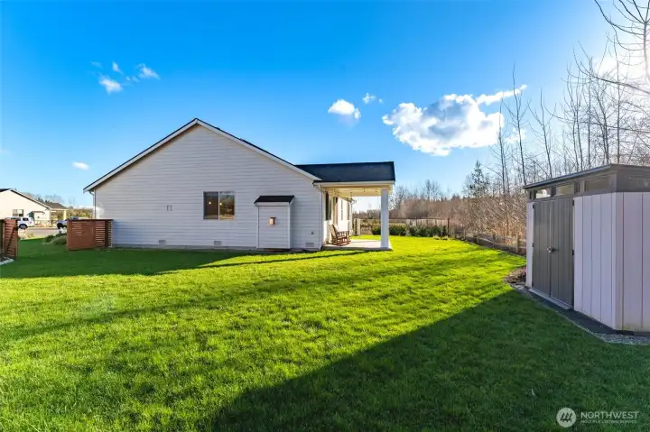 Custom cedar fencing with gated access surrounds the oversized backyard, featuring open green space, raised garden beds, landscaped borders, a composite storage shed, and a rear elevation view highlighting the covered patio and seamless indoor-outdoor living.