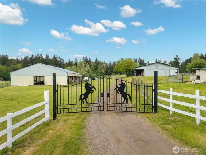 A grand equestrian-inspired gated entrance introduces this luxury home on private acreage, framed by white fencing, open grounds, and expansive outbuildings.