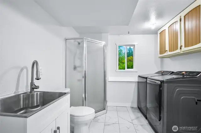 Lower-level laundry room with tile floors, utility sink, washer/dryer with shower and natural light.