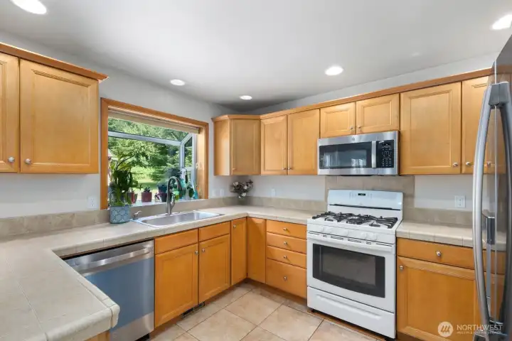 U-shaped kitchen layout with generous prep space, tile counters, recessed lighting, and a large window above the sink bringing in natural light and backyard views.