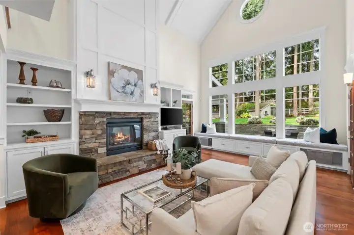 Sunlight pours into the soaring great room, where a stone-clad fireplace serves as a grounding centerpiece against expansive windows. This luminous heart of the home seamlessly connects the refined interior to the lush Fox Island landscape just beyond the glass.