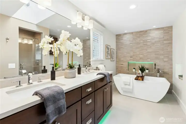 Spa-inspired primary bath featuring dual quartz vanities, oversized mirror, designer lighting, heated-style ambiance lighting, and a stunning freestanding soaking tub framed by textured stone tile accents.