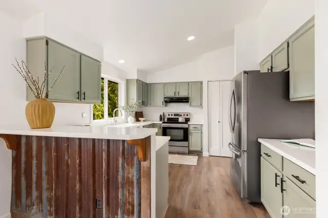 The kitchen is both welcoming and practical with sage green cabinetry, stainless steel appliances, and a custom corrugated metal breakfast bar—bringing personality and function together in one unique package.