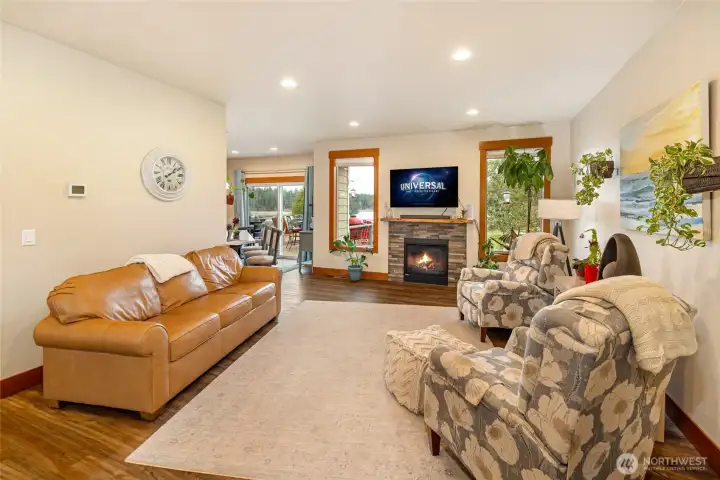 Bright and welcoming living room featuring warm wood floors, a cozy stone-trimmed fireplace, and large windows that frame peaceful views of the surrounding landscape. Perfect for relaxing evenings or entertaining guests.