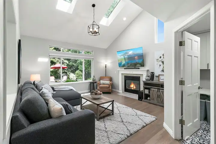 Bright living space with vaulted ceiling, fireplace, large windows, natural light, hardwood flooring, and backyard views.