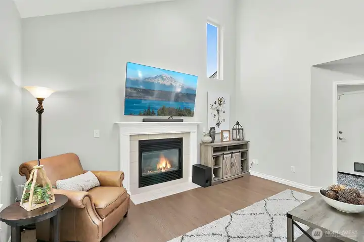 Bright living space with vaulted ceiling, fireplace, large windows, natural light, hardwood flooring, and backyard views.
