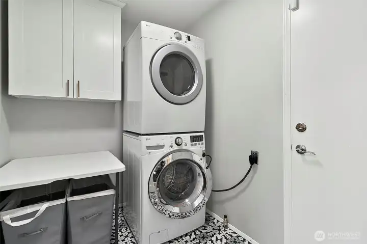 Laundry room with stacked washer and dryer, built-in cabinet storage, folding counter, patterned tile flooring, and exterior door access.