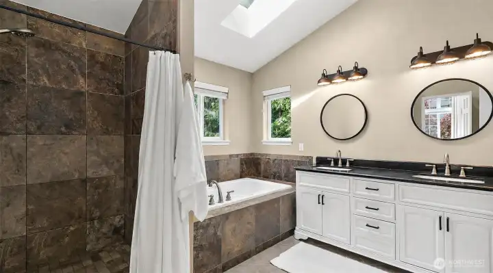 Primary 5 piece bathroom with skylight, soaking tub, walk-in tile shower, dual sink vanity, stone tile accents, and natural light.