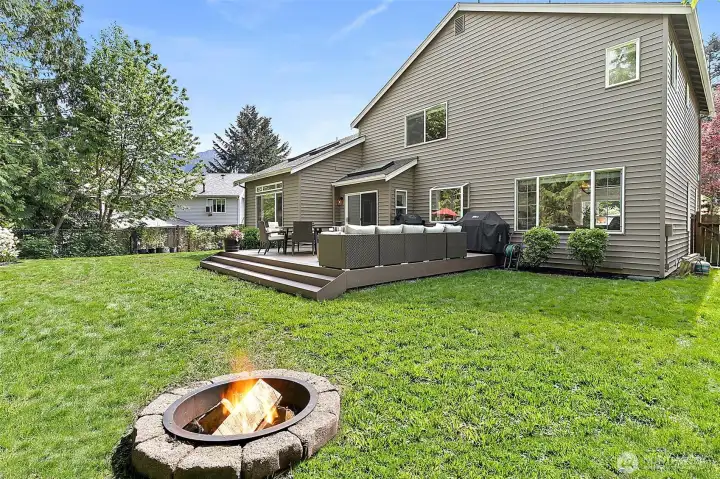 Fully fenced backyard with new deck, outdoor dining and lounge areas, fire pit, lawn space, mature landscaping, and greenbelt backdrop.